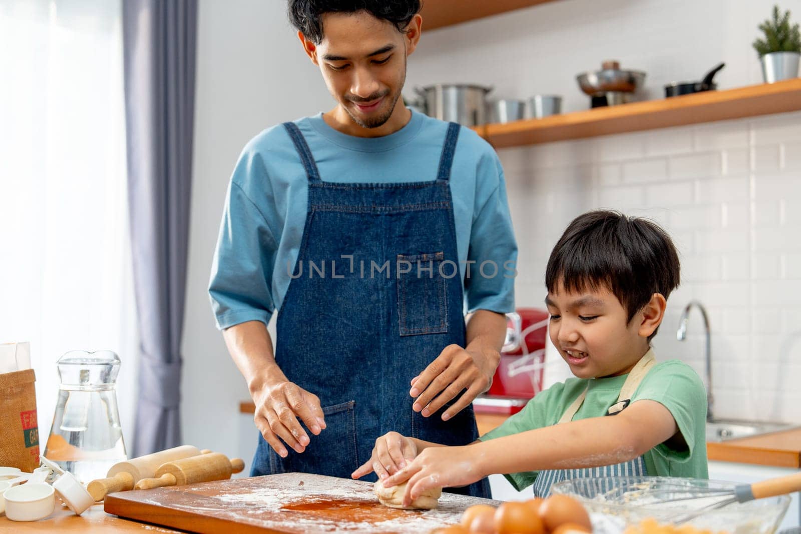 Asian father stand beside his son during teaching the boy to threshing flour for making bakery in kitchen of their house.