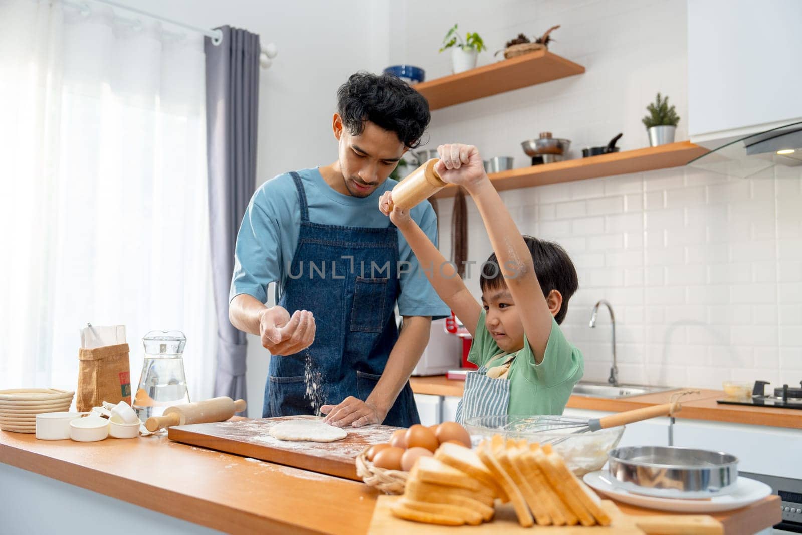 Young Asian father pour cooking flour on wooden tray during teaching his son who hold rolling pin for threshing flour for making bakery in kitchen of their house.