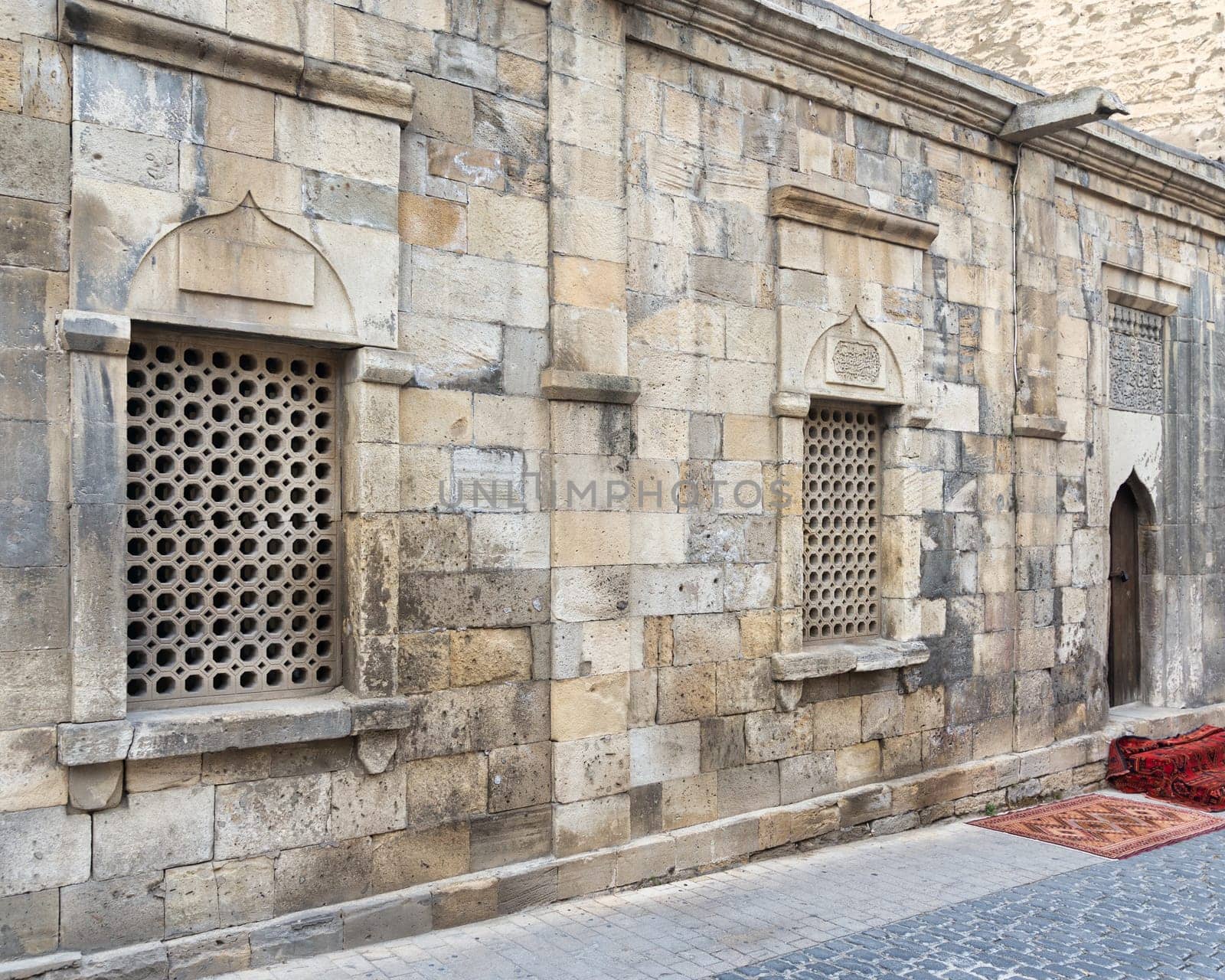 The narrow streets of Icherisheher in Baku, Azerbaijan, featuring the intricate stonework of a historic building. The warm afternoon light highlights the unique lattice windows and textured walls