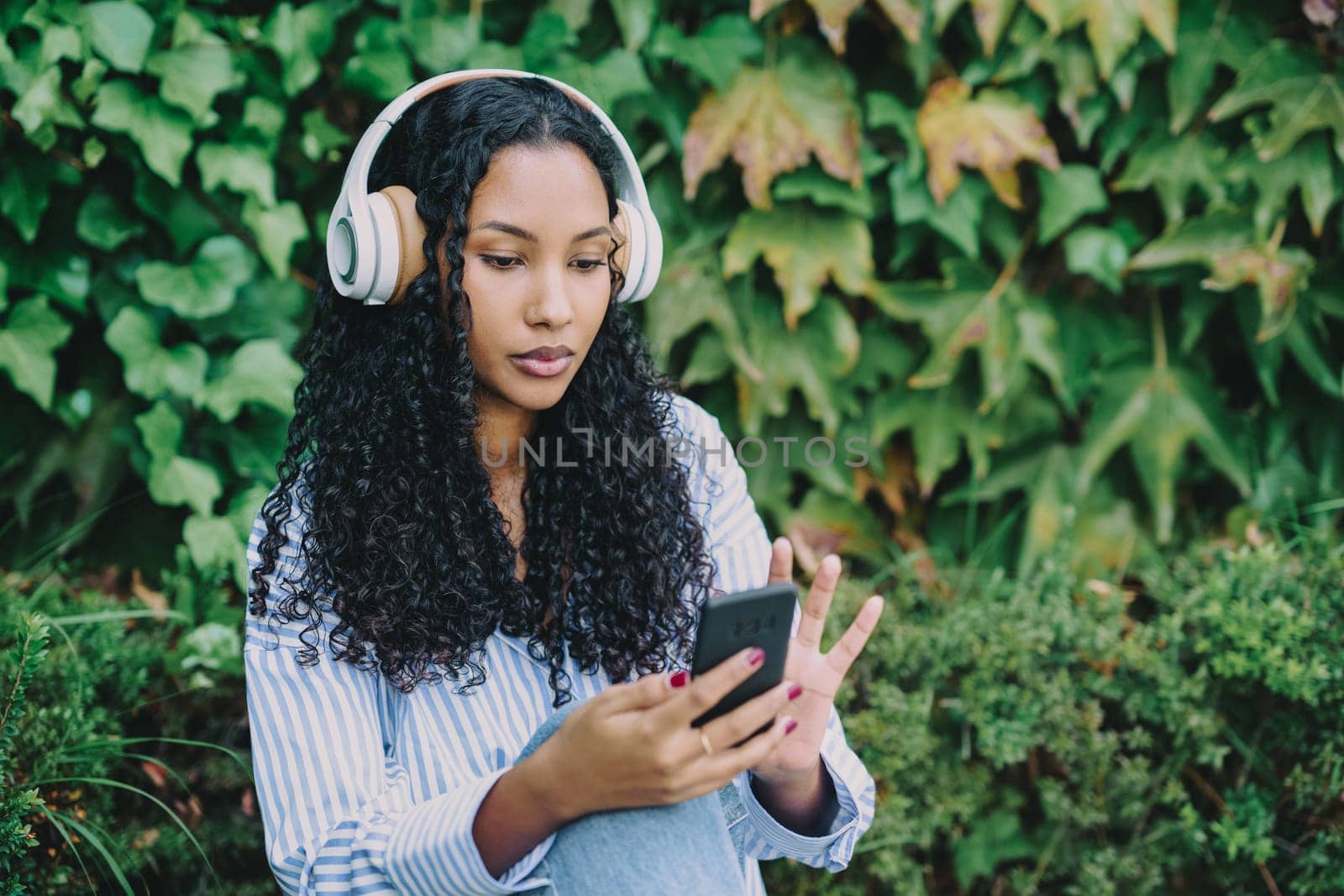 A young woman with beautiful curly hair is sitting outdoors in a peaceful setting, wearing stylish headphones and busily using her smartphone while being surrounded by vibrant greenery all around her