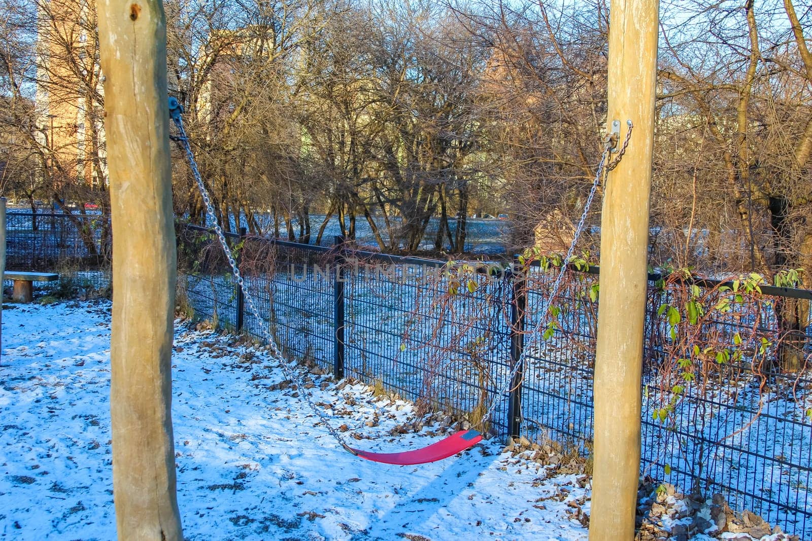 A swingset sits in a snowy park surrounded by leafless trees and a river, evoking a serene winter atmosphere.