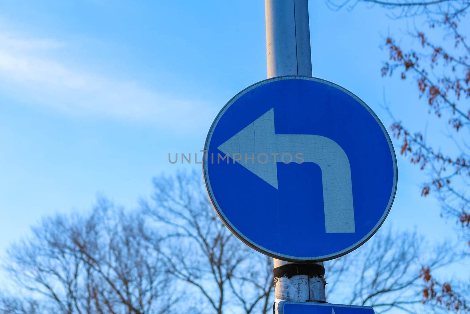 A clear blue road sign indicates a left turn in an urban area, surrounded by bare trees under a bright sky.