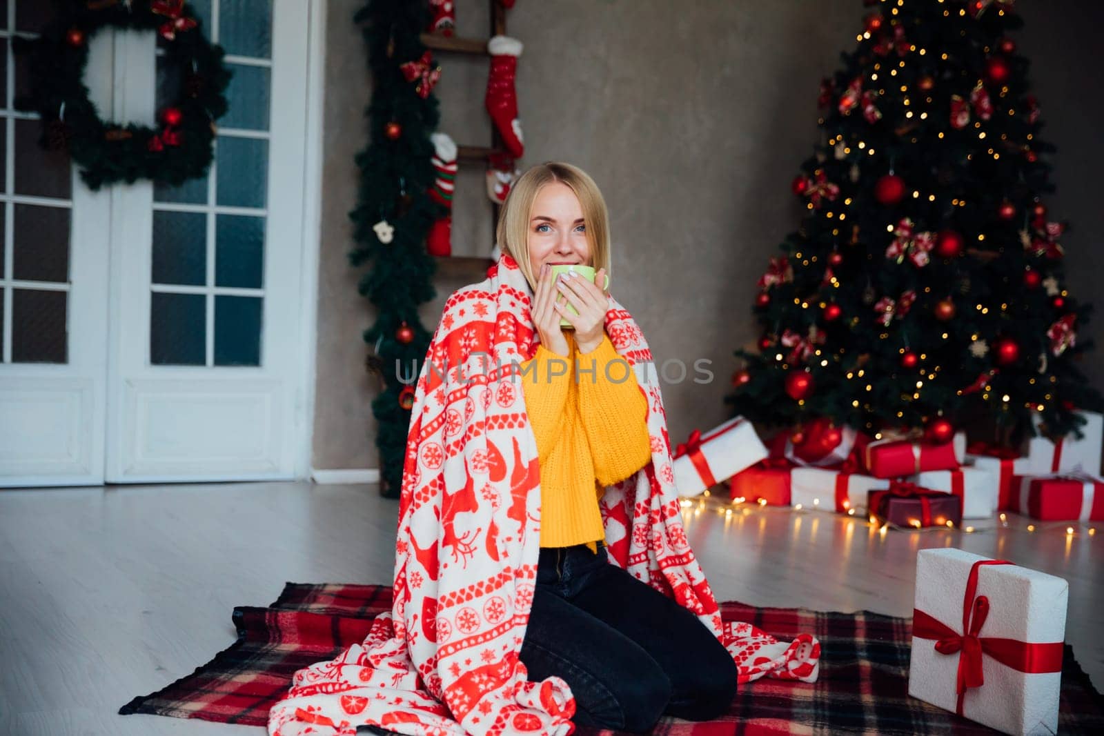 blonde woman in a blanket sitting with a mug in a room with a Christmas tree Holiday Christmas