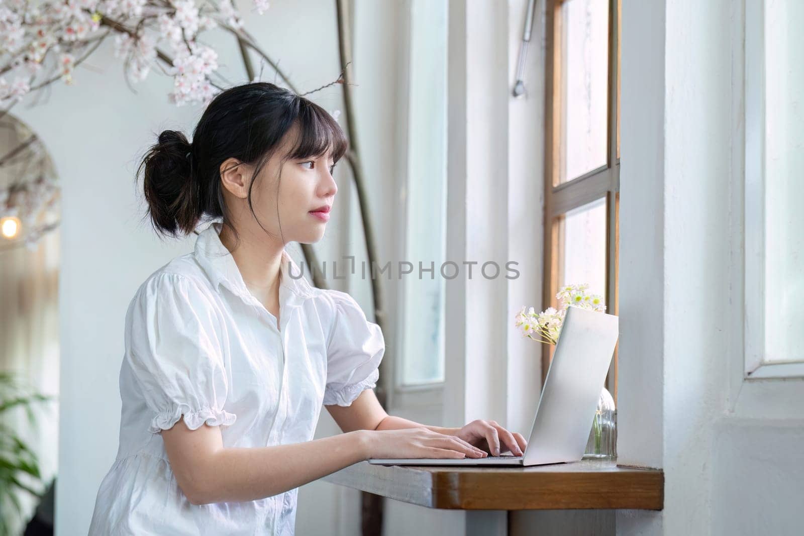Asian female student studying online in living room with laptop doing online research for a class subject Make notes for essay homework. online education concept by wichayada