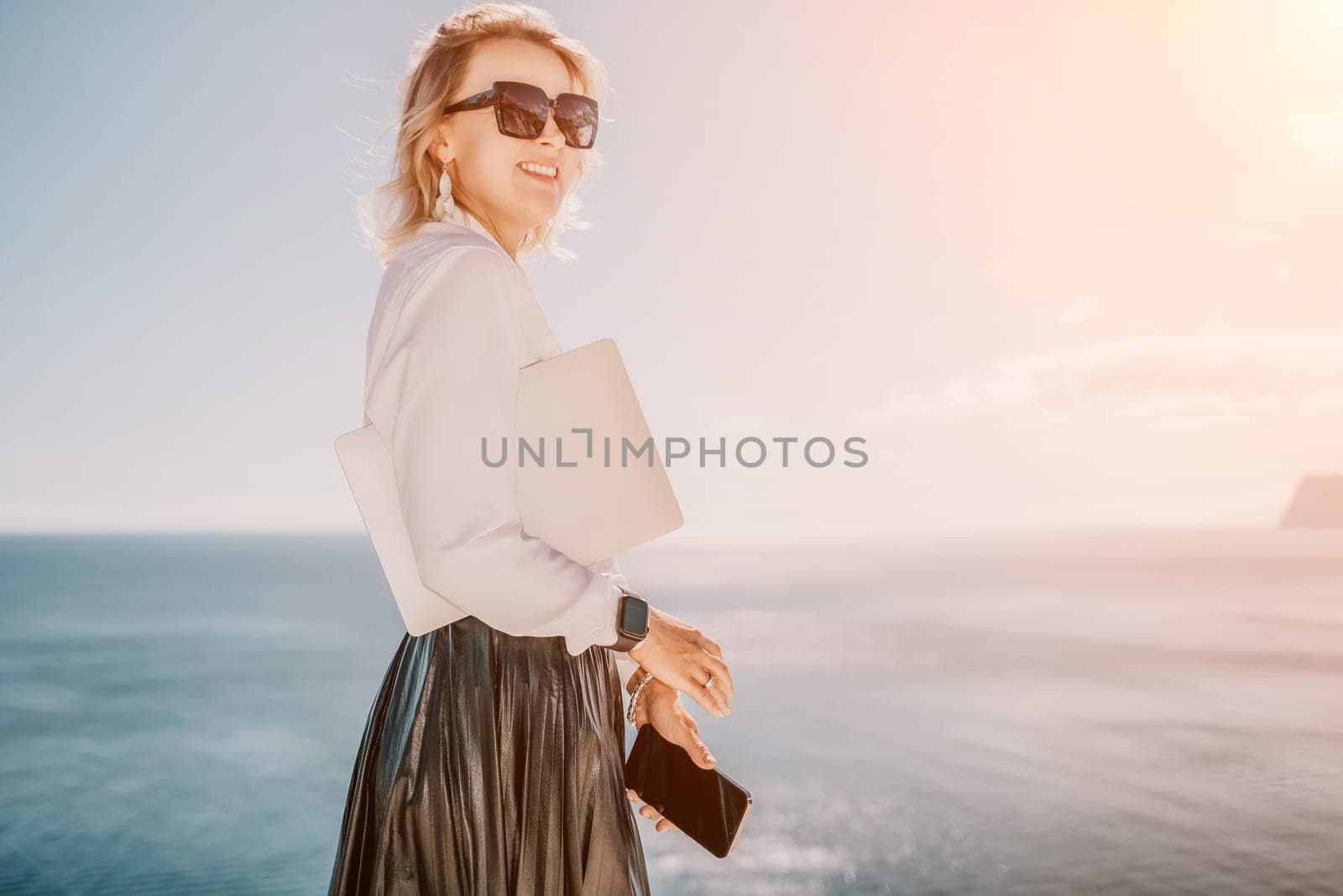 Digital nomad, Business woman working on laptop by the sea. Pretty lady typing on computer by the sea at sunset, makes a business transaction online from a distance. Freelance remote work on vacation