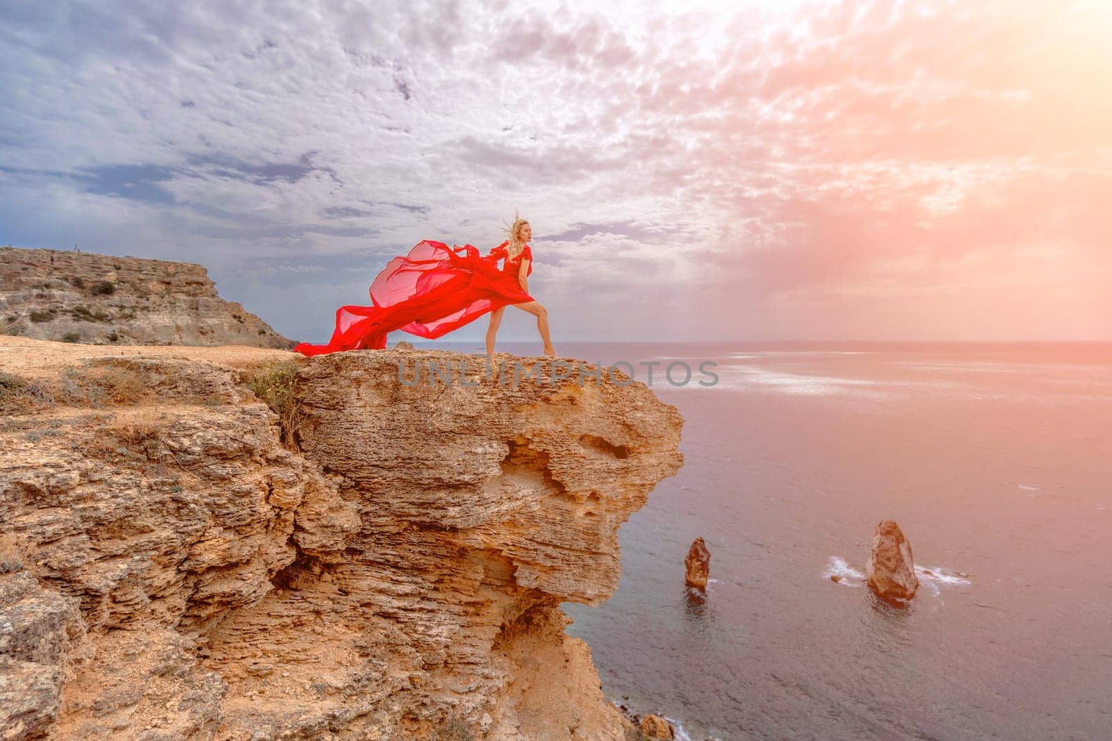 woman red silk dress stands by the ocean, with mountains in the background, as her dress sways in the breeze. by Matiunina