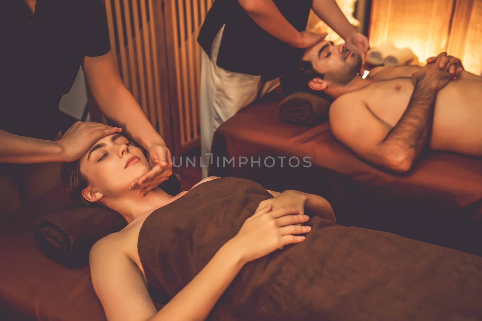 Couple customer enjoying relaxing anti-stress head massage and pampering facial beauty skin recreation leisure in warm candle lighting ambient salon spa in luxury resort or hotel. Quiescent