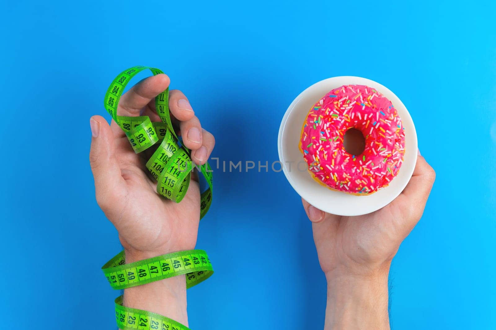 Male hand holding a pink donut on a blue background. Choosing between a donut and a measuring tape. Health and healthy lifestyle or unhealthy sweet food.