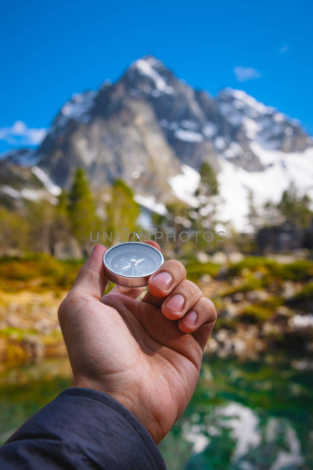 A man with a compass in his hand in the high mountains near a clear lake. Travel concept. Landscape photography. A man traveler is looking for direction with a compass on the coast near a lake in the mountains, first person view.