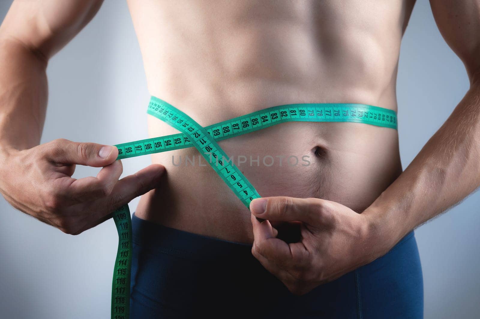 A man of thin build with muscle definition and six-pack abs measures the circumference. of his waist with a centimeter and a length measuring tape