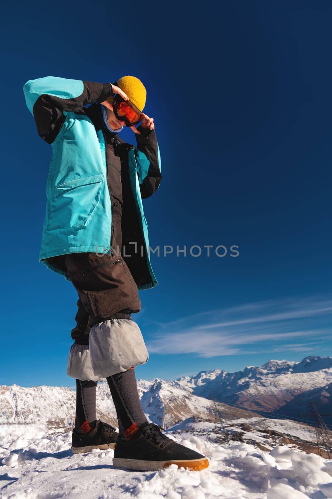 A stylish fashionable skier in a hat and sneakers adjusts his ski goggles against a background of mountains. Vertical frame with copy space.