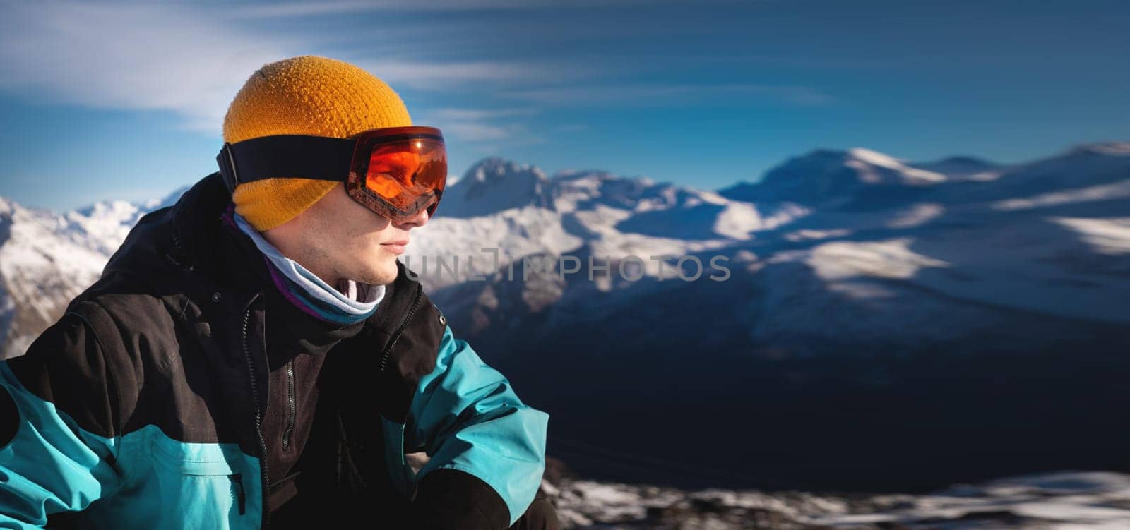 young man in ski goggles on a snowy mountain on a sunny day. close-up of a man looking to the side, enjoying the mountain scenery, relaxing after skiing on a sunny day. Snowboard holiday concept.