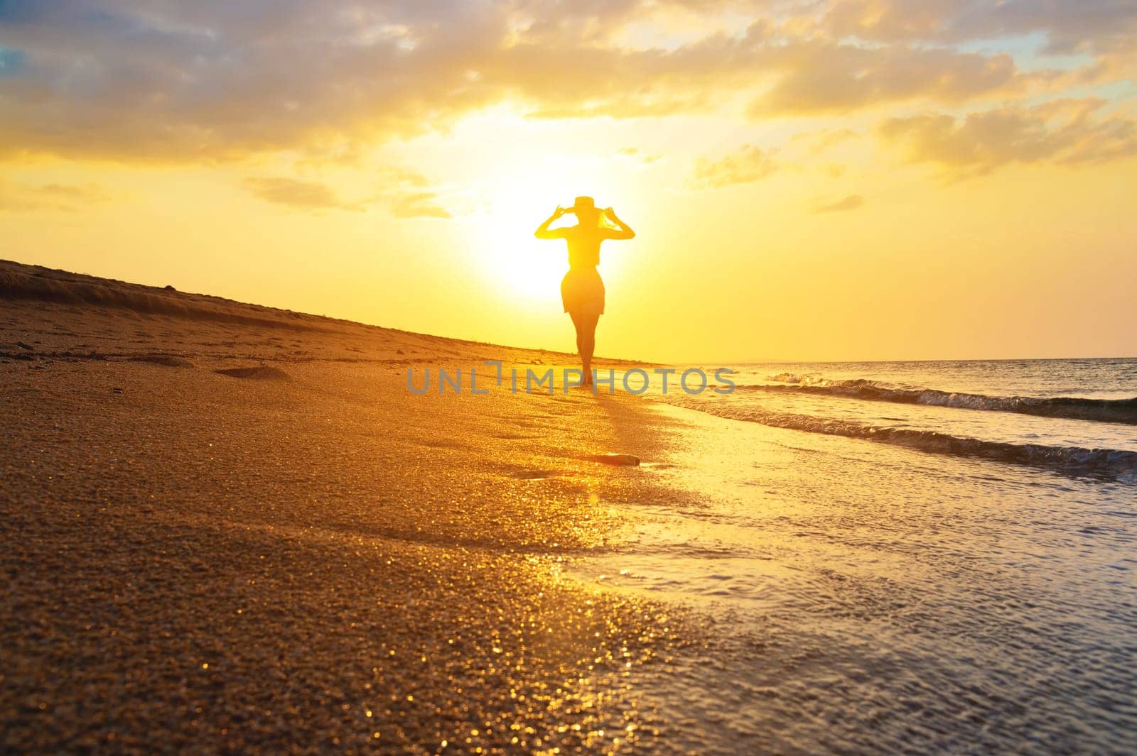 Happy slim young woman carefree walking on the beach with calm waves at sunset. Silhouette landscape with a slender woman and a golden sandy beach.