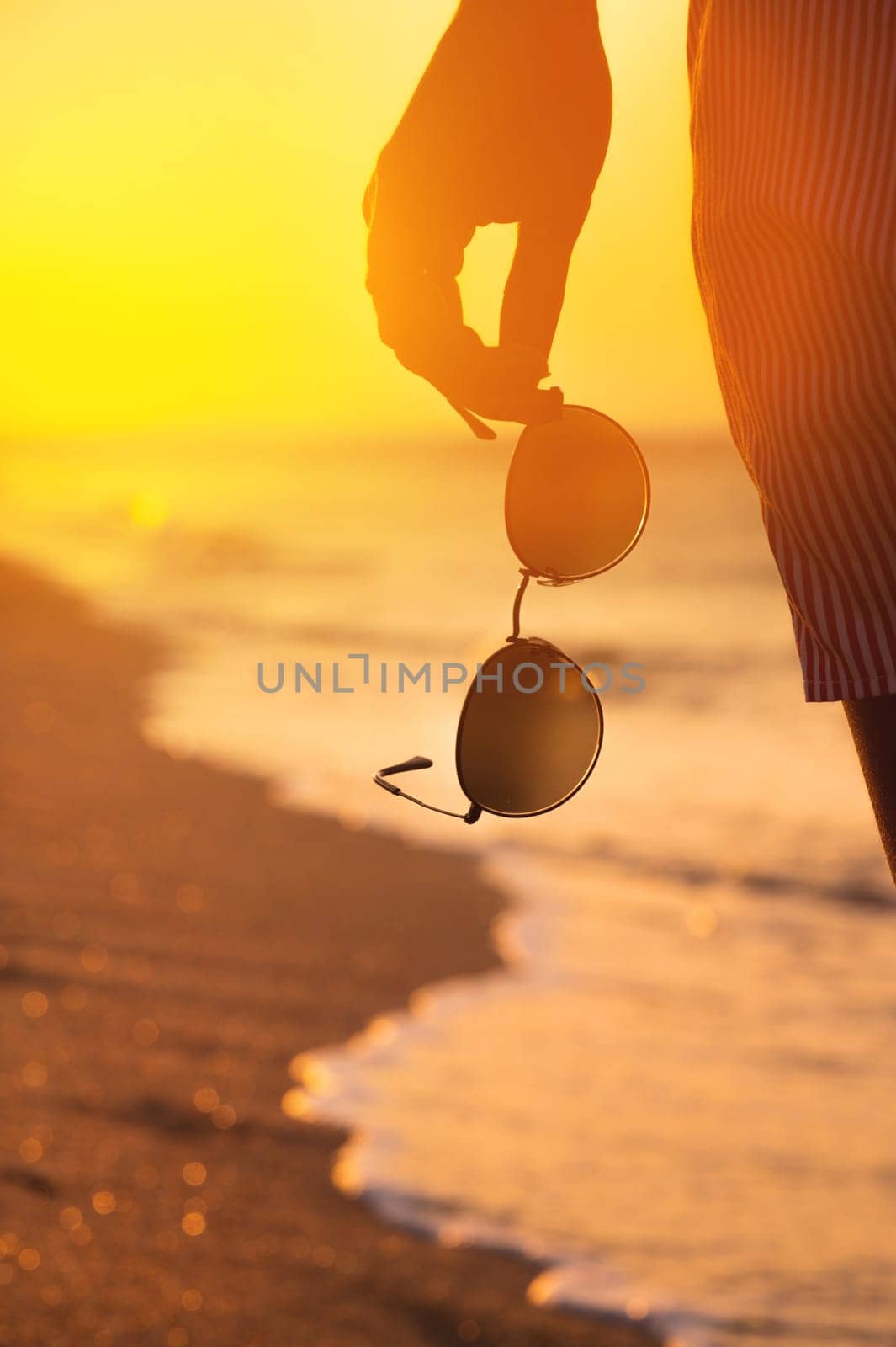 Close-up of a woman's hand holding stylish round sunglasses at sunset. Summer atmosphere near the sea coast, a woman on a walk holds glasses in her hand. Vertical shot.