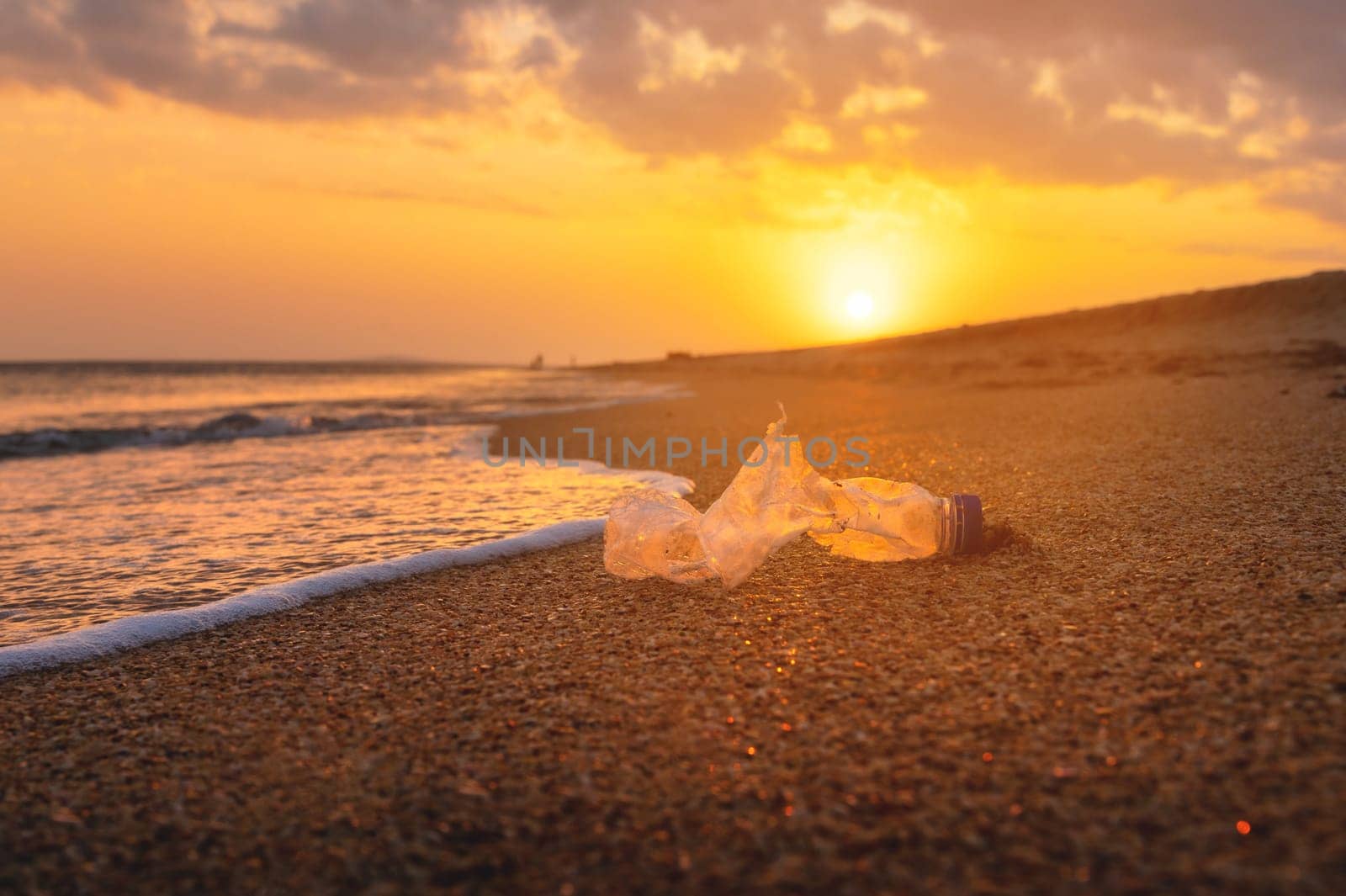 Close-up of environmental and sea pollution concept. Ocean pollution from plastic waste. An old torn bottle lies on wet golden sand at sunset.