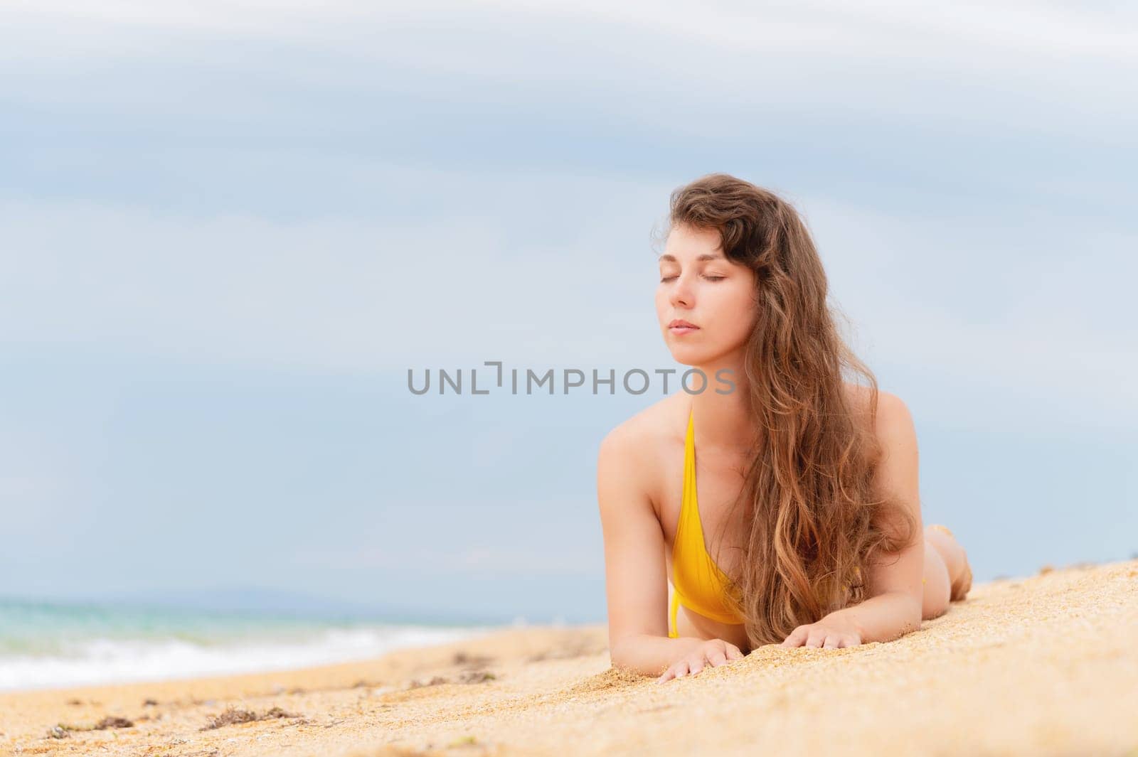 Beautiful woman with perfect body lies on sandy beach, wearing yellow bikini, sunbathing at beach resort, enjoying summer vacation.