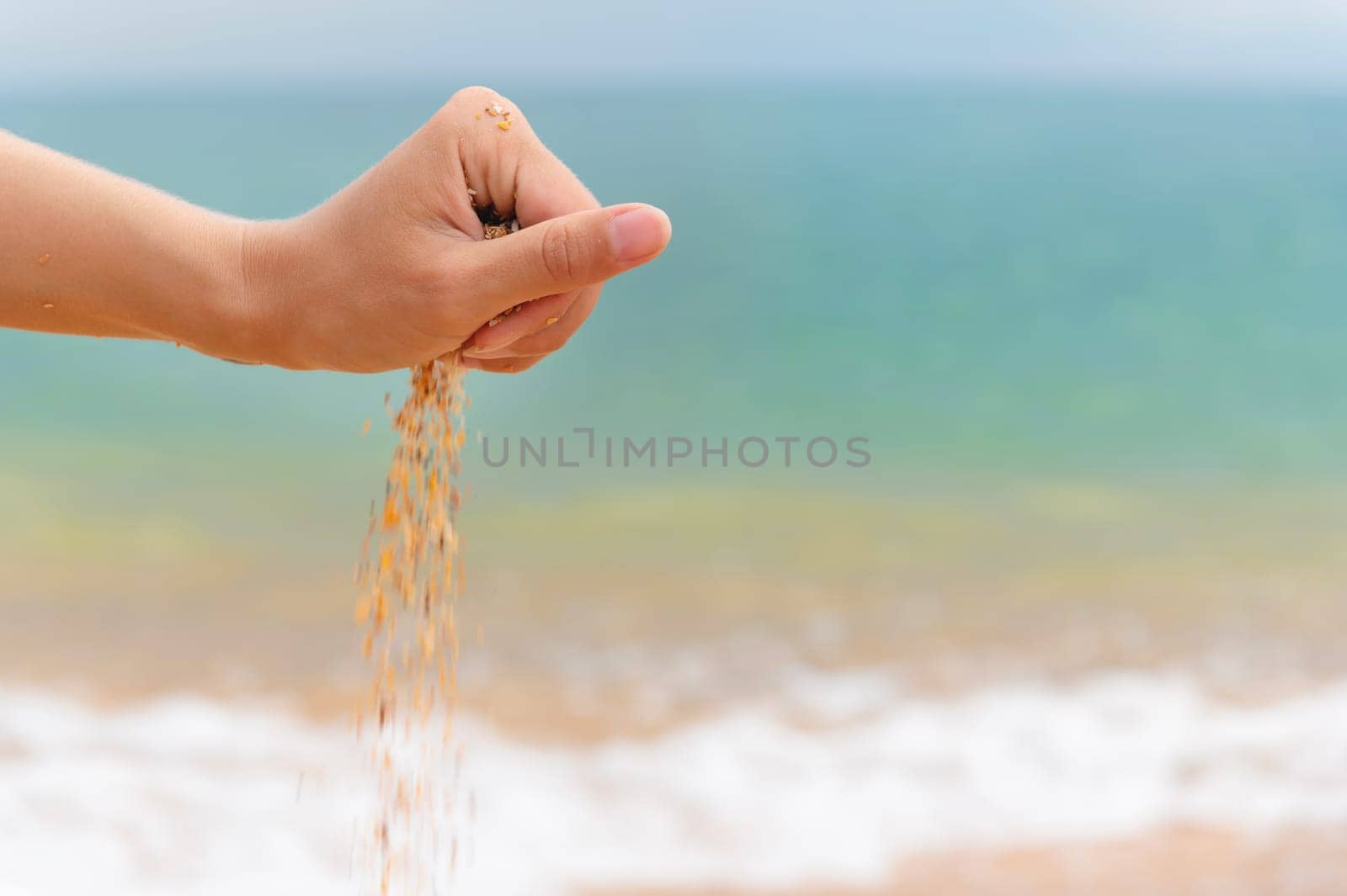 Close-up of a woman's hand releasing falling sand. Sand flows through the hand against the backdrop of the turquoise sea. Summer beach holiday concept.