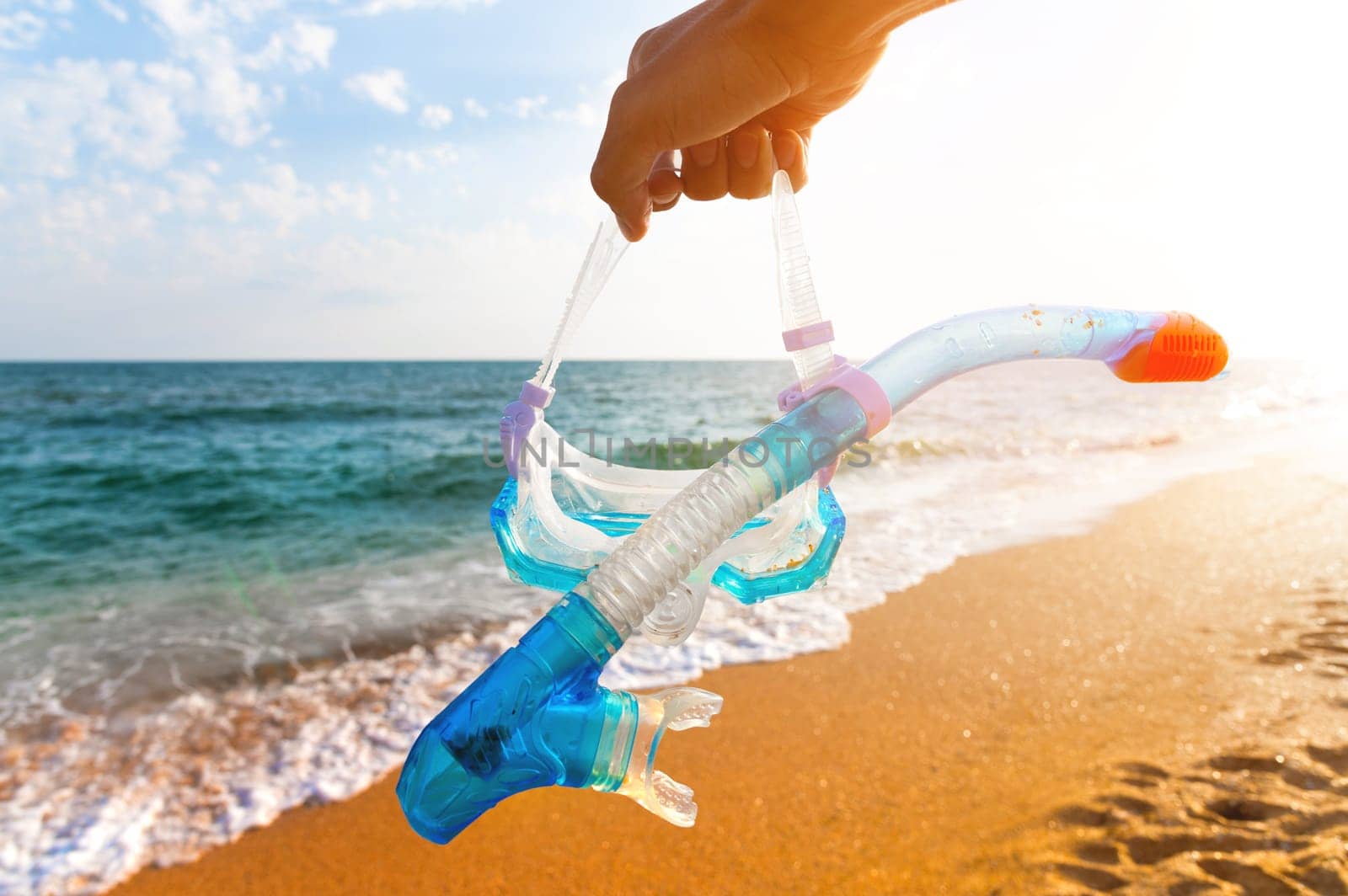 A hand holds a snorkeling mask and snorkel over the ocean water. Summer holiday, bright sun on the beach