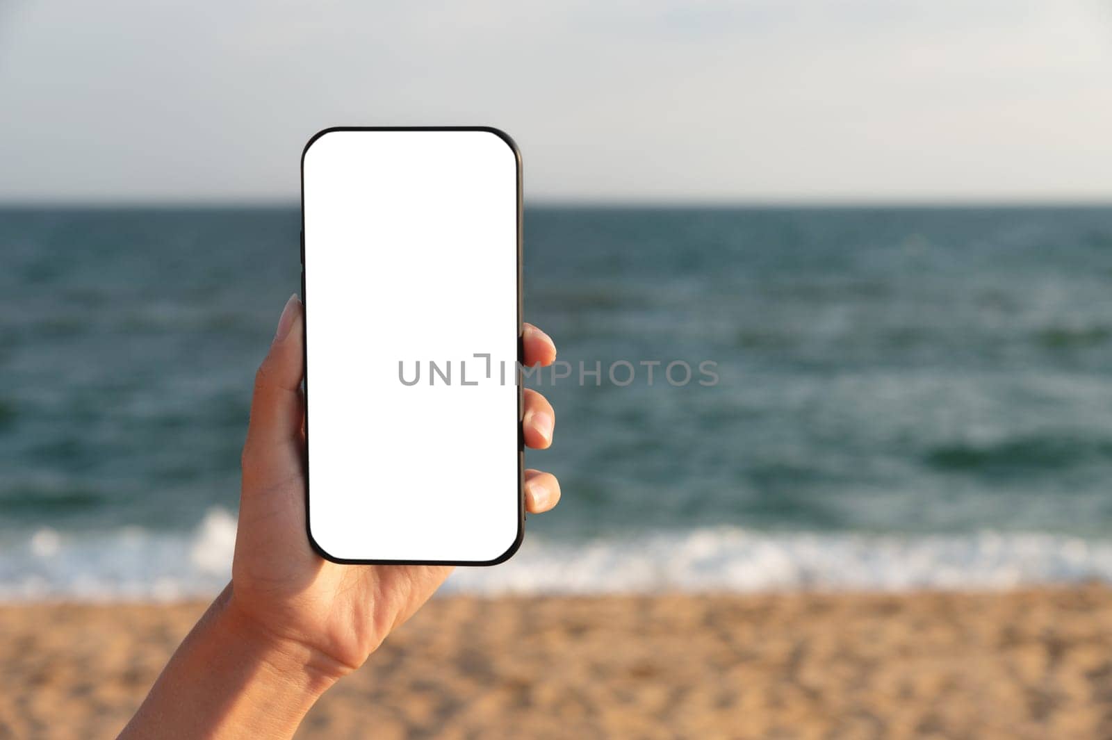 Mockup image of a woman's hand holding a white mobile phone screen against a background of blue sky, sea and beach. place for your advertising.