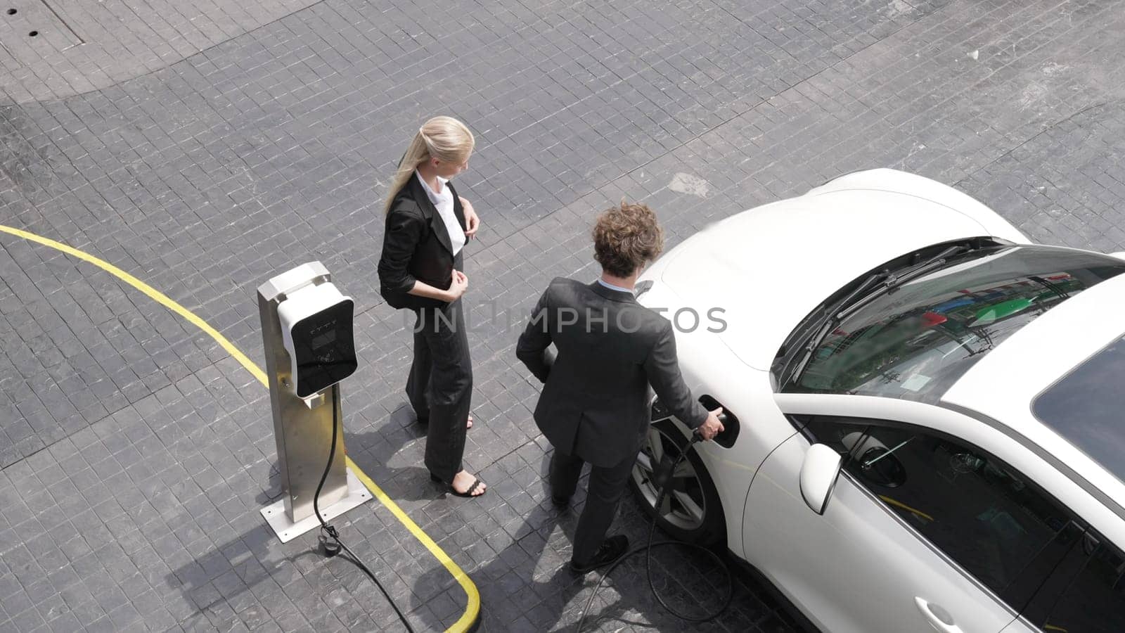 Progressive businessman and businesswoman leaning on electric car connected to charging station before driving around city center. Eco friendly rechargeable car powered by clean energy.