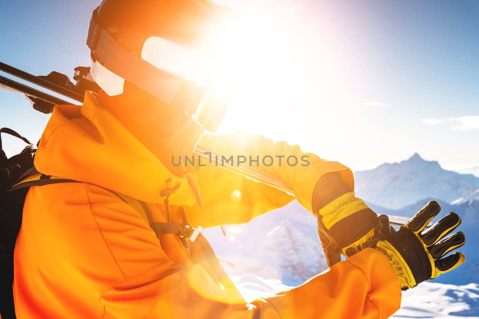 A young unrecognizable man holds skis on his shoulder in bright clothes, gloves and a ski mask, standing against a background of mountains and a clear sky, with a bright sun