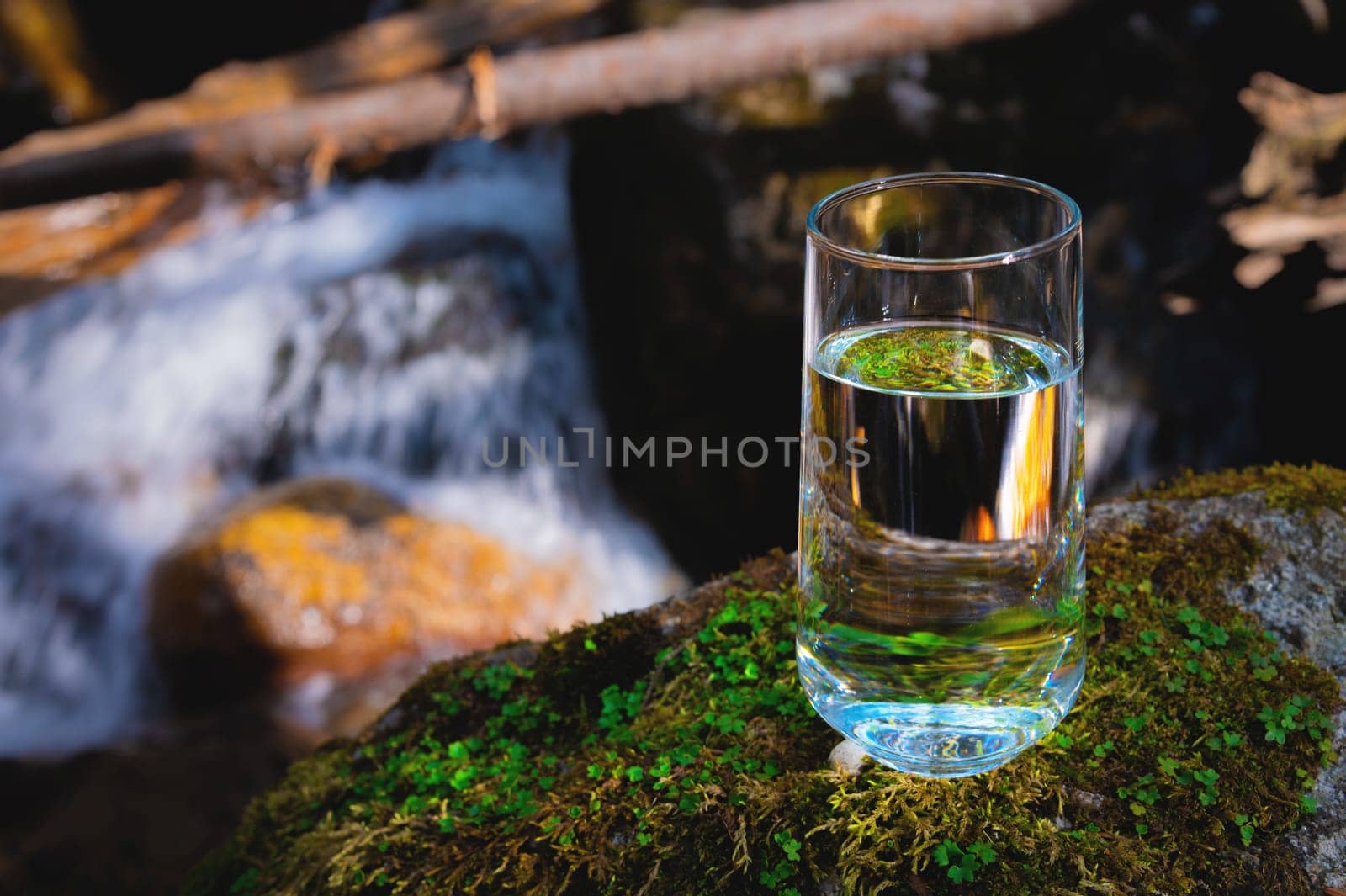 Clear water in a clear glass against a background of green moss with a mountain river in the background. Healthy food and environmentally friendly natural water by yanik88