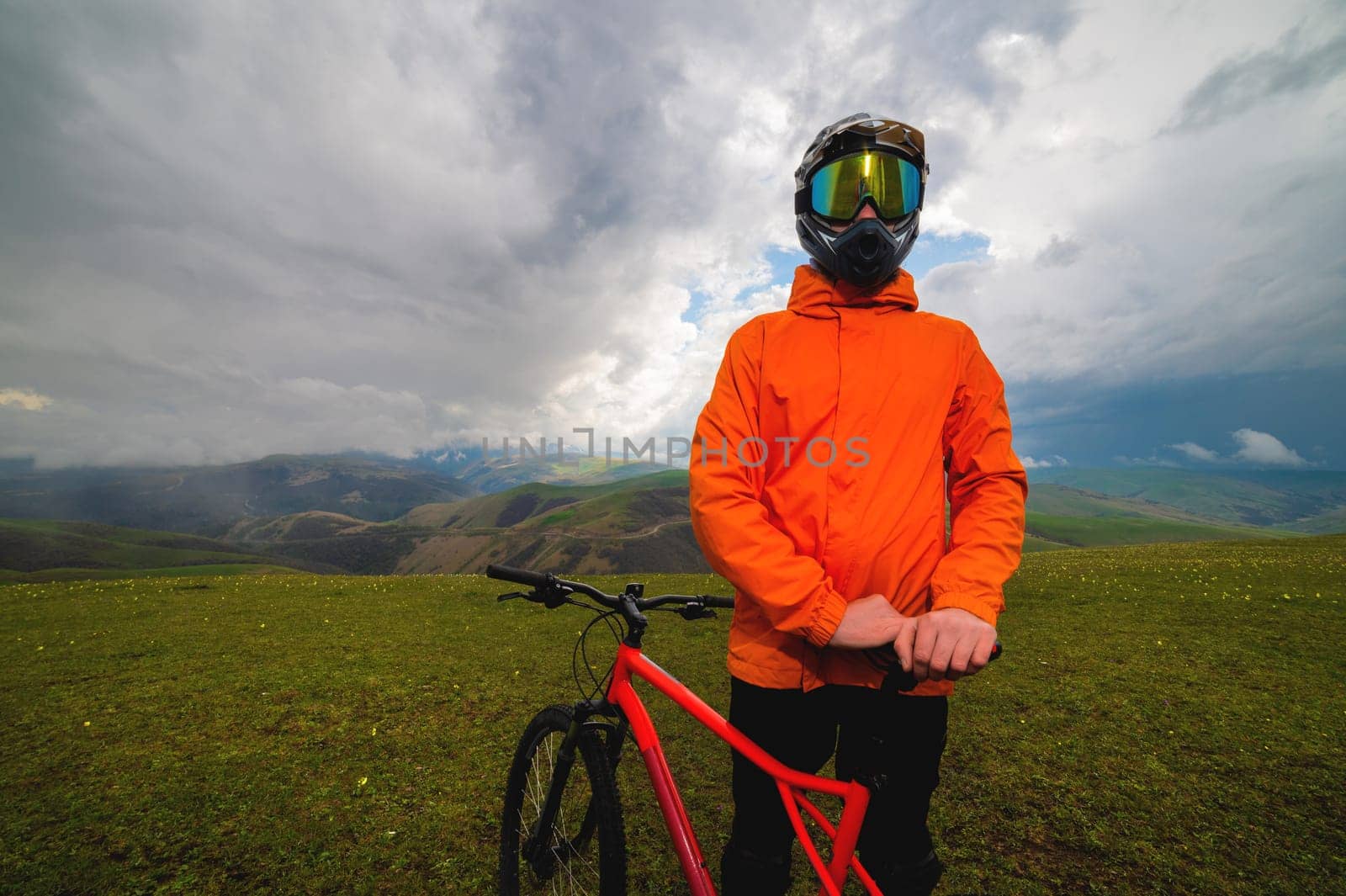 A cyclist in an orange bright jacket and a protective helmet stands and holds a bicycle in the mountains, outside the city, off the road in summer in cloudy weather.
