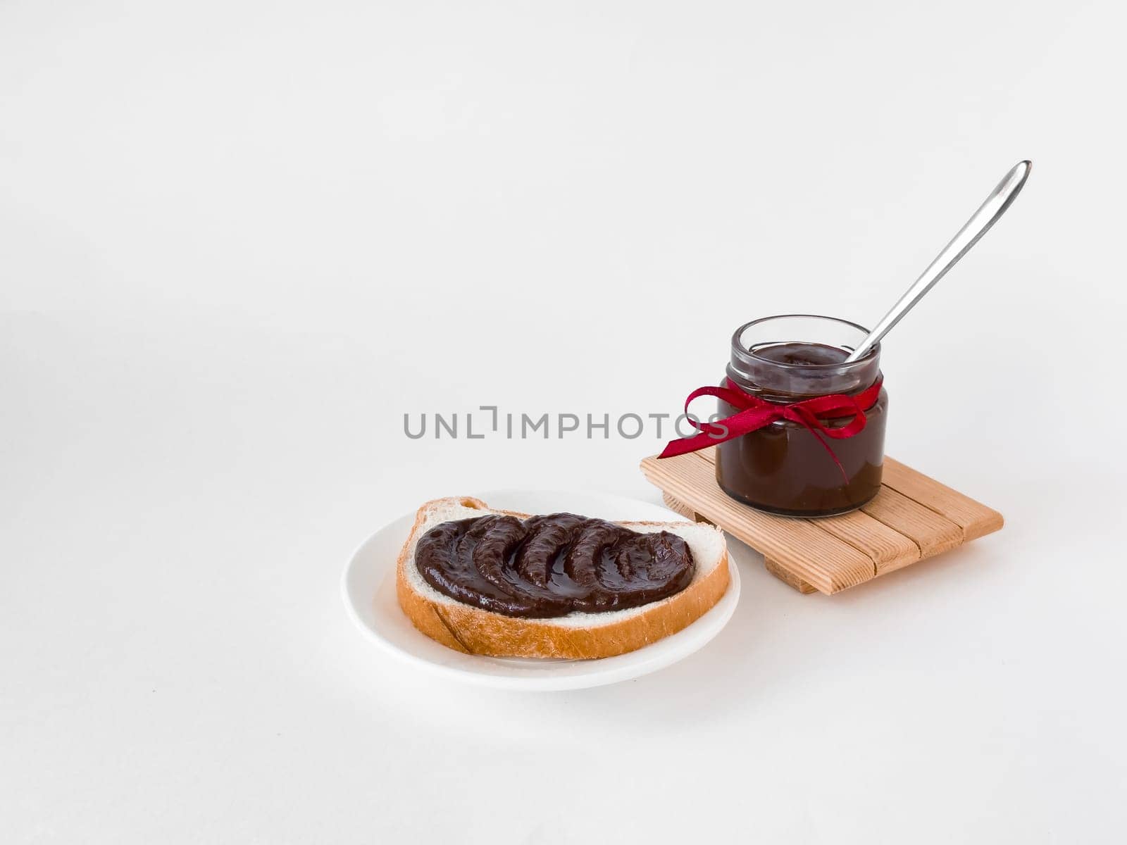 Chocolate spread on rustic bread with jar and spoon on white background.