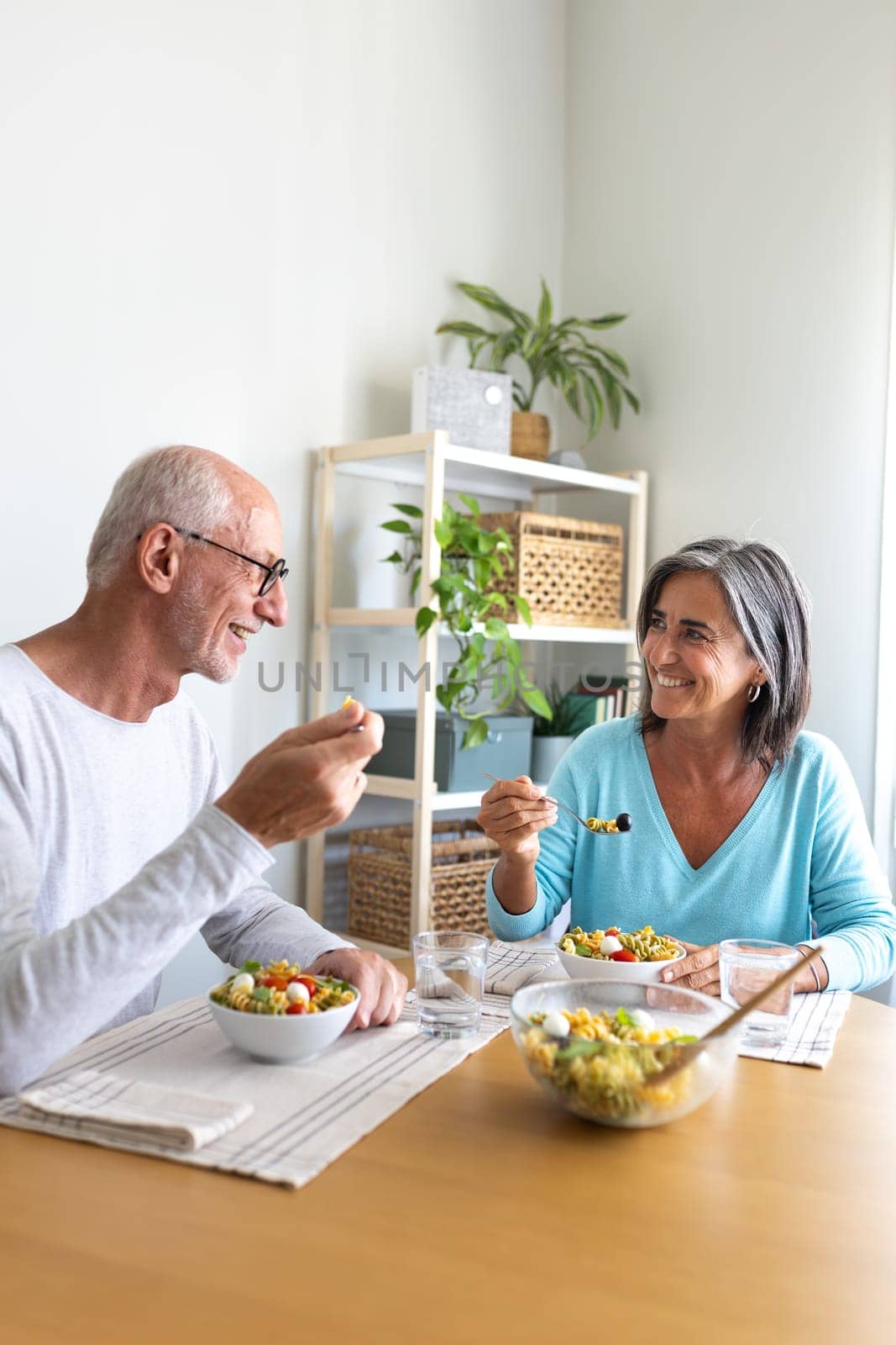 Mature married couple enjoying pasta salad for lunch. Couple looking at each other smiling while eating. Vertical. Lifestyle concept.