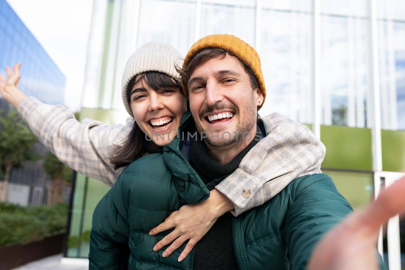 Cheerful man giving piggyback ride to girlfriend while taking selfie in city