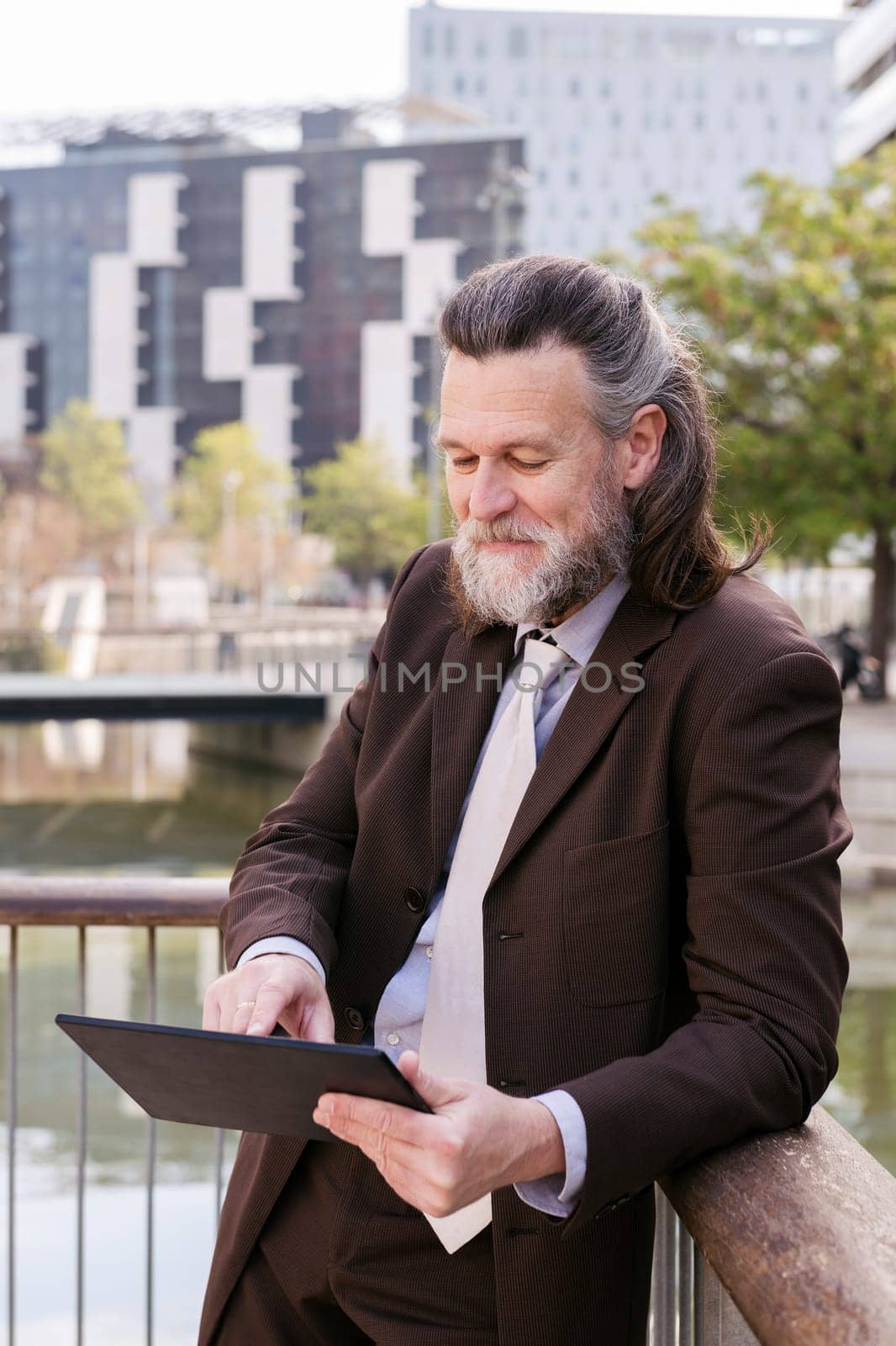 positive middle age male with beard in formal outfit browsing tablet while standing at city, concept of business and mature man lifestyle, copyspace for text