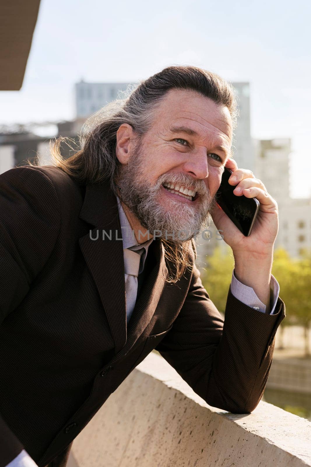 smiling middle age male with beard and gray hair in classy suit discussing business issues on phone conversation against modern city buildings, concept of business and mature man lifestyle