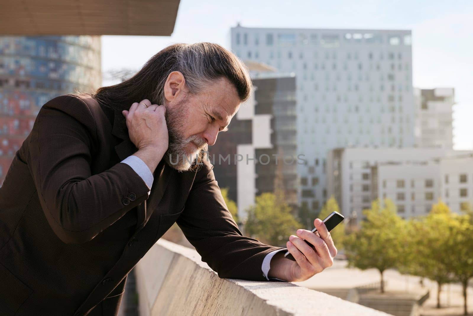 concerned mature male with beard in classy suit using mobile phone while standing against modern city buildings, concept of technology of communication and middle age man lifestyle, copyspace for text