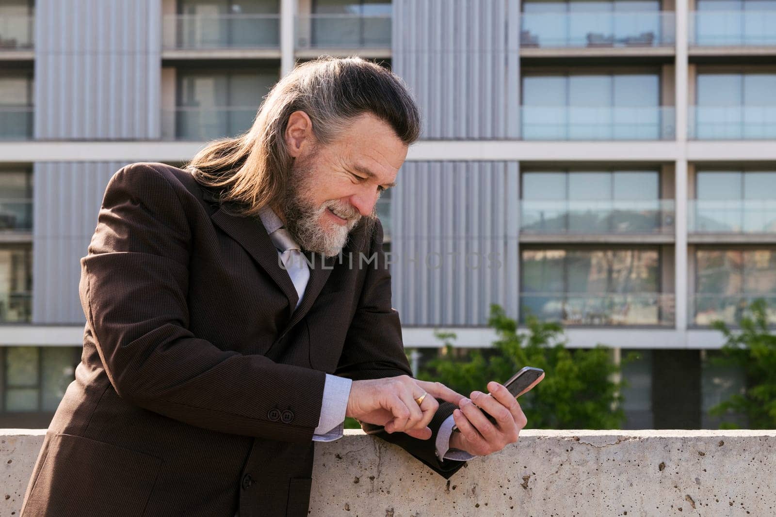 smiling middle age male with beard and gray hair in classy suit browsing on smartphone against modern city buildings, concept of business and mature man lifestyle, copyspace for text