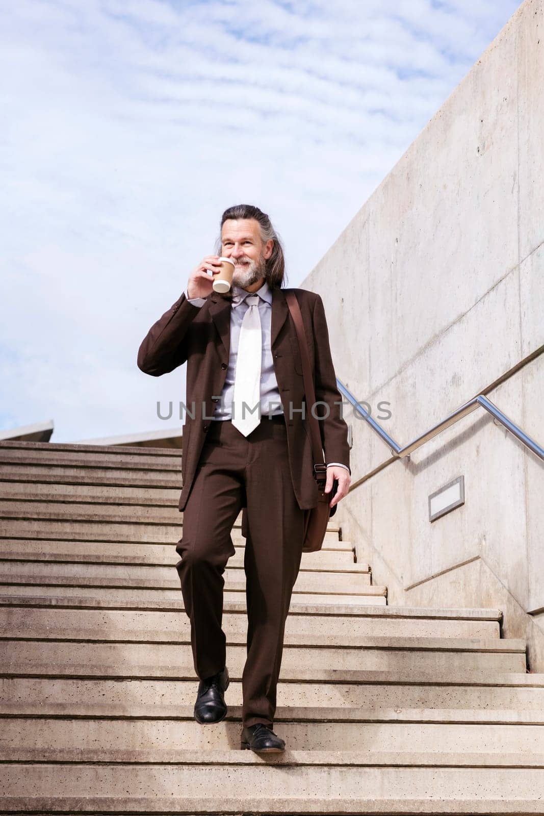 stylish mature businessman in classy suit with briefcase and takeaway coffee walking down urban stairs in city, concept of business and middle age man lifestyle