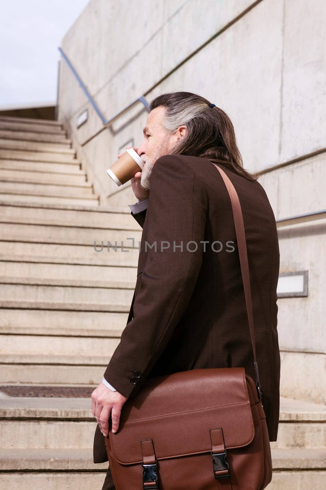 rear view of confident mature male executive manager in elegant suit with bag and takeaway coffee walking on urban stairs, concept of business and middle age man lifestyle, copyspace for text