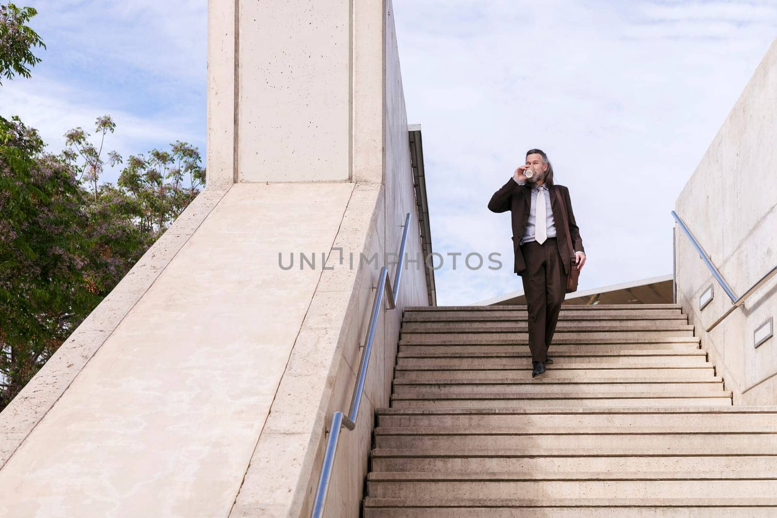 stylish mature businessman in classy suit with briefcase and takeaway coffee walking down urban stairs in city, concept of business and middle age man lifestyle, copyspace for text