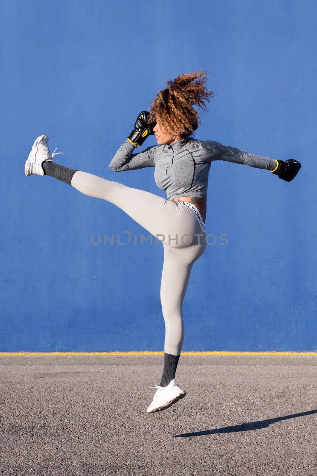 latin female fighter in sportswear and boxing gloves jumping with raised leg while exercising on blue background, concept of strength and healthy lifestyle