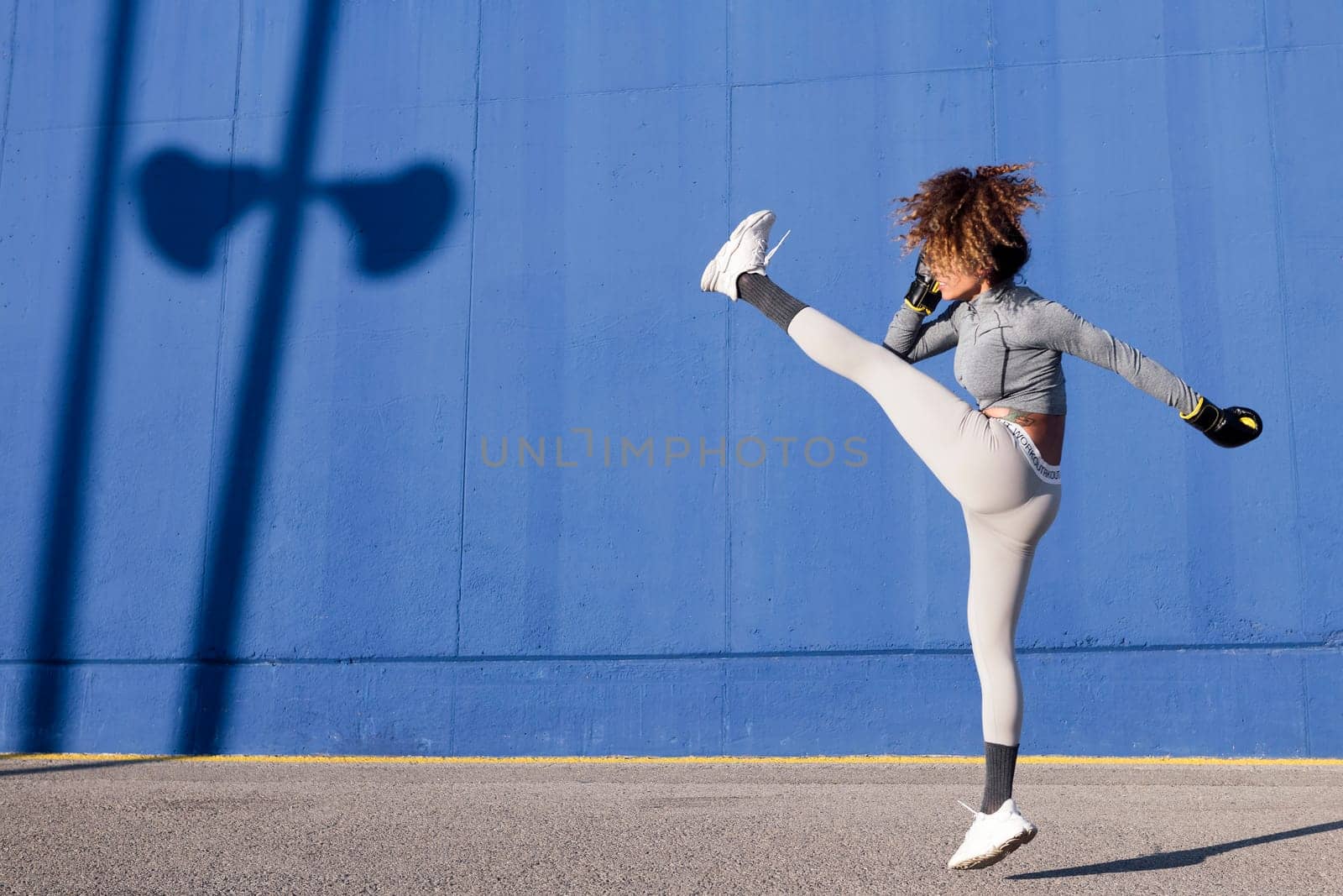 latin female fighter in sportswear and boxing gloves jumping with raised leg while exercising on blue background, concept of strength and healthy lifestyle, copyspace for text