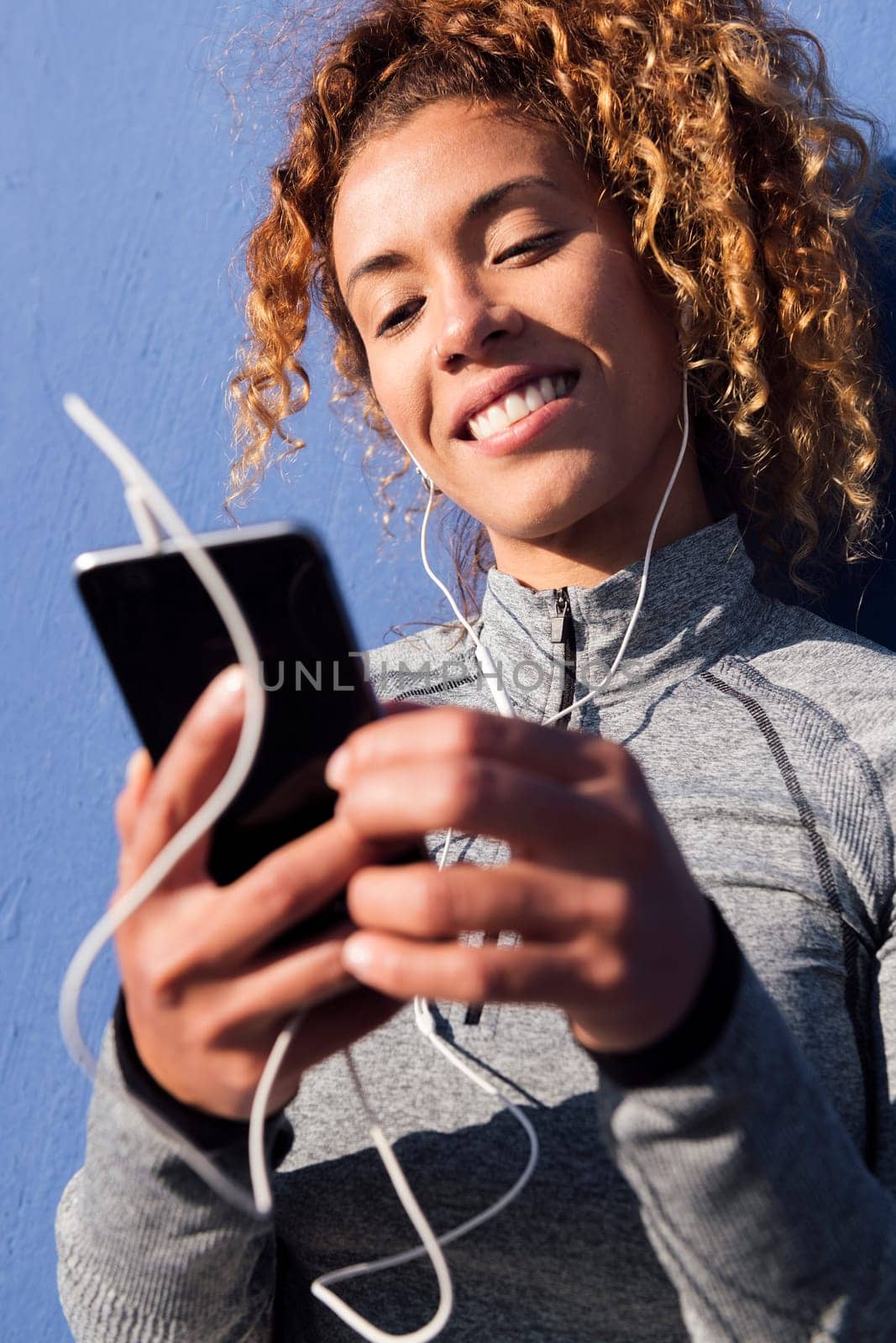 latin female athlete in earphones smiling happy while using mobile phone during break from workout on blue background, concept of urban sport and technology of communication