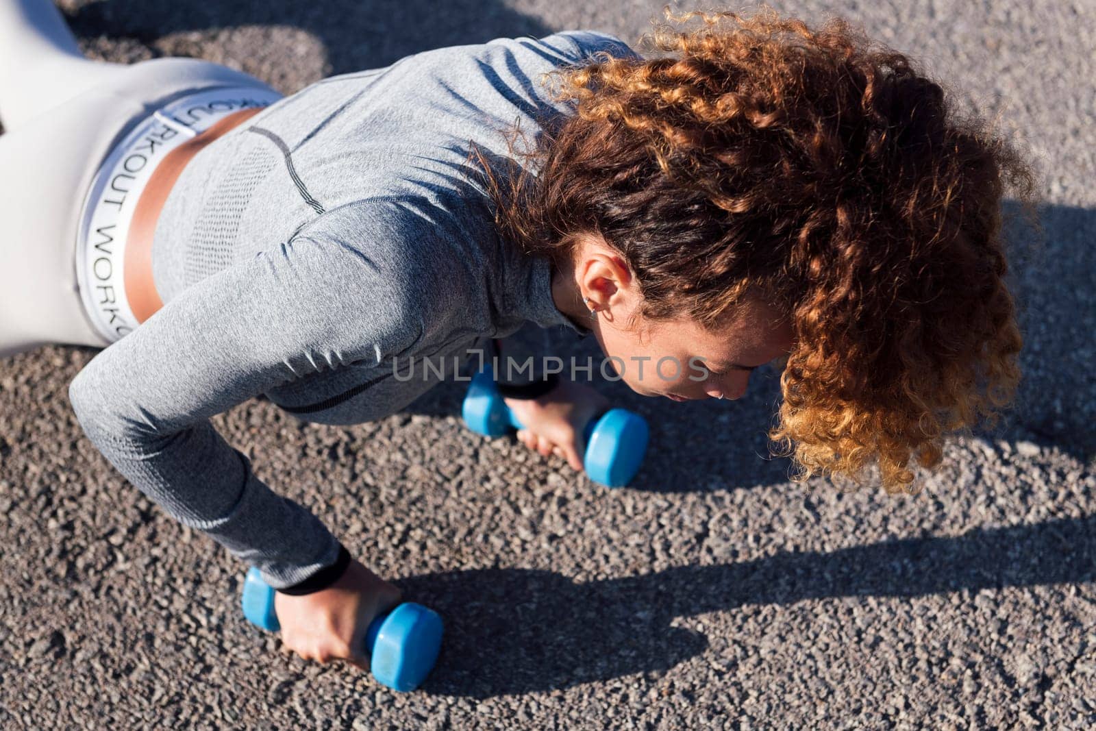 strong female athlete doing planks with dumbbells during training on pavement, concept of urban sport and healthy lifestyle