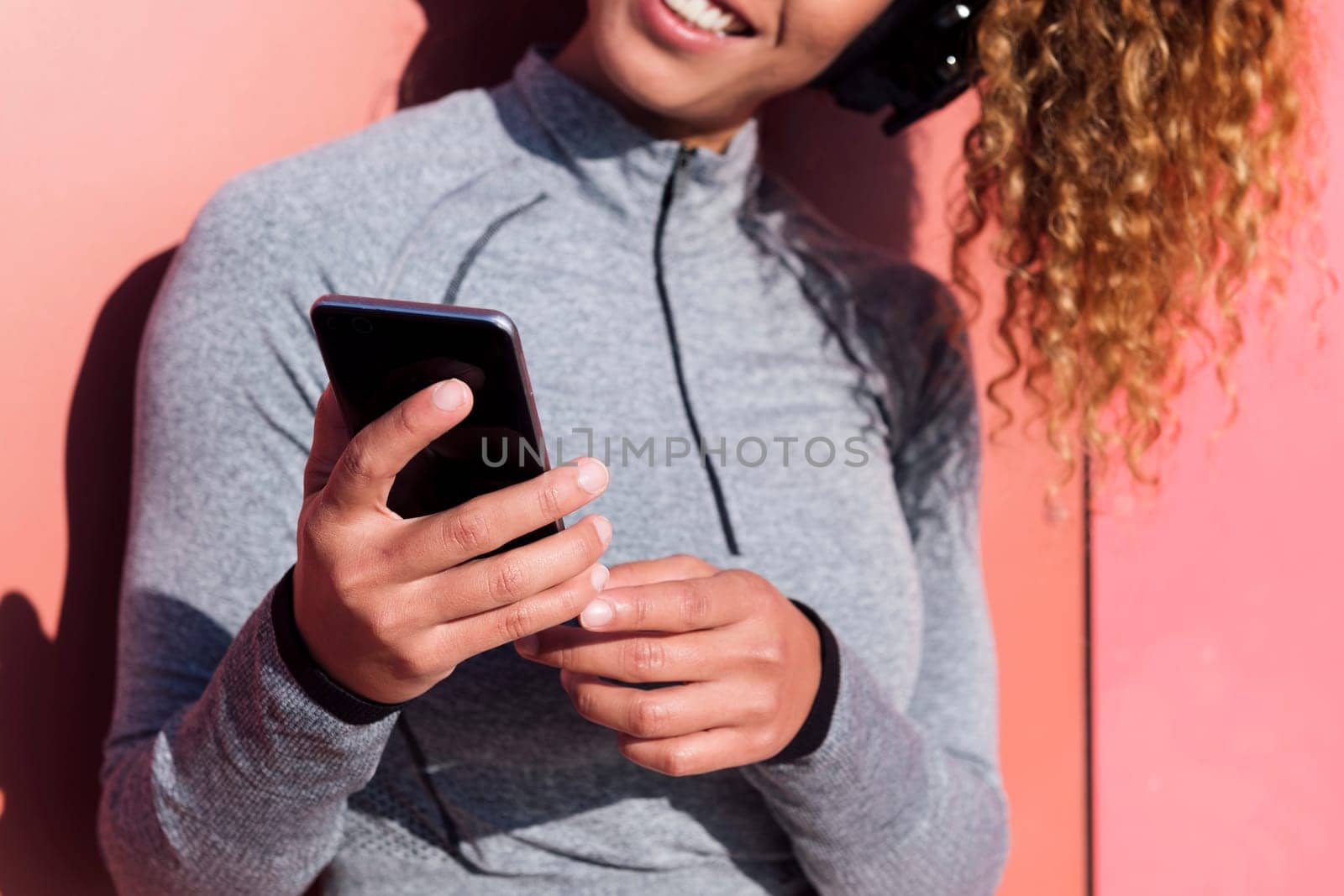 close up of the hands of a woman with mobile phone listening to music from headphones against pink wall, concept of technology of communication and urban lifestyle, selective focus on the phone