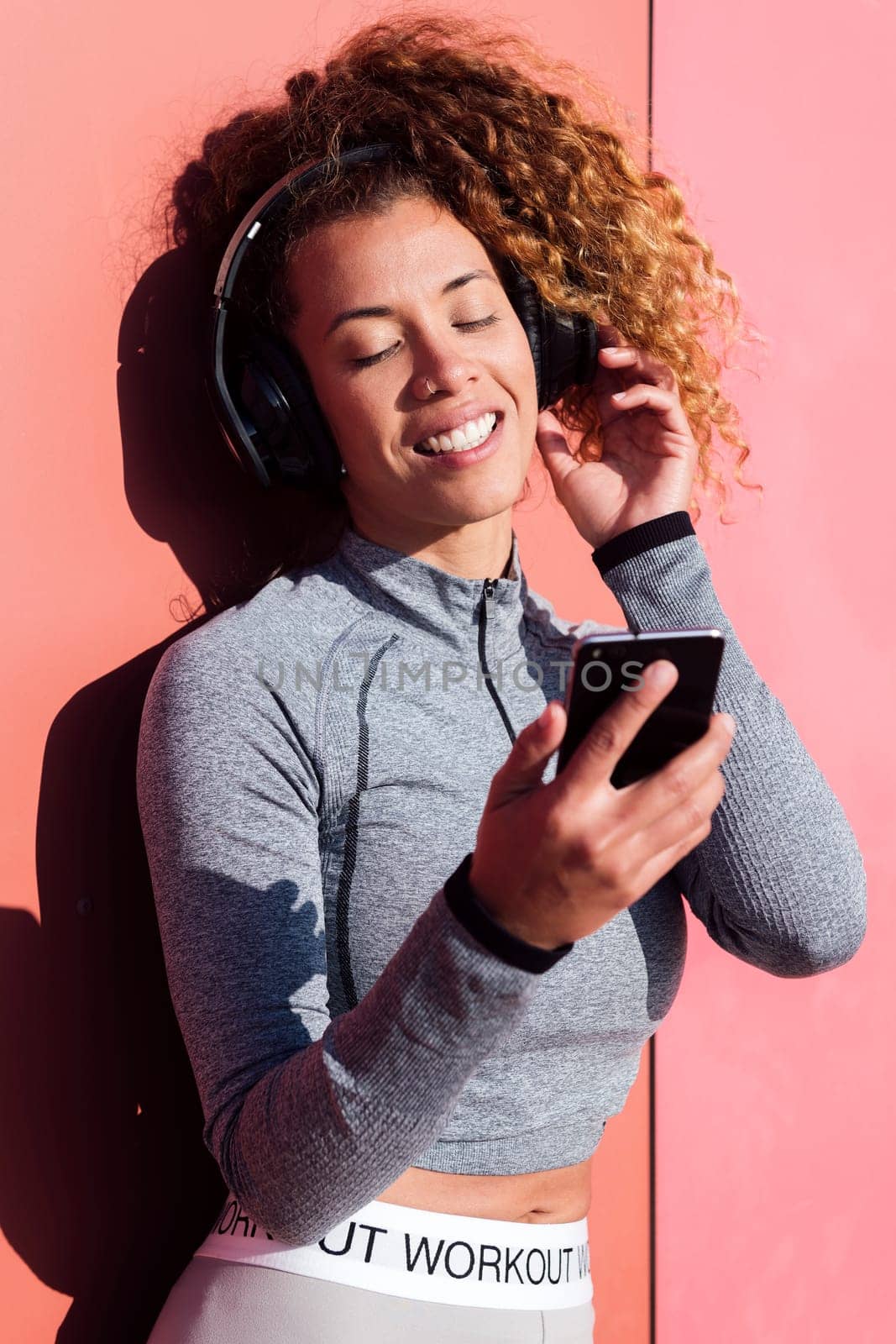 cheerful sportswoman with mobile phone listening to music from headphones against pink wall, concept of technology of communication and urban lifestyle