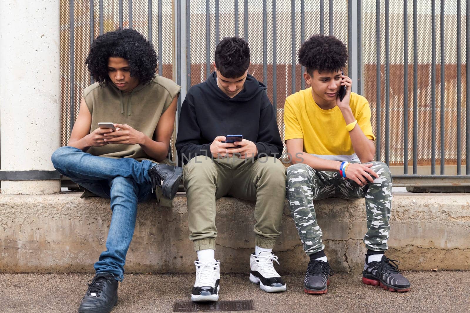 three serious multiracial teen friends browsing Internet on mobile phones sitting against metal fence and looking at screen, concept of youth and technology of communication