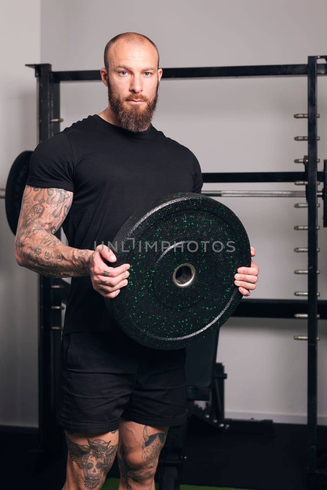 powerful sportsman in sportswear standing with black weight plate in hands and looking at camera during workout in gym, concept of fitness and active lifestyle