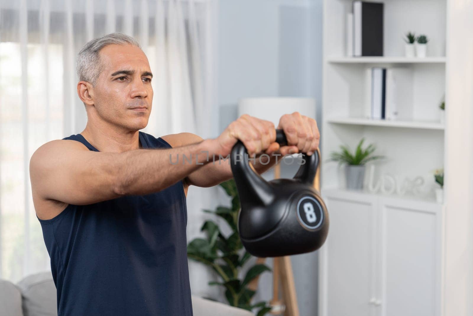 Athletic and sporty senior man engaging in body workout routine with lifting kettlebell at home as concept of healthy fit body with body weight lifestyle after retirement. Clout