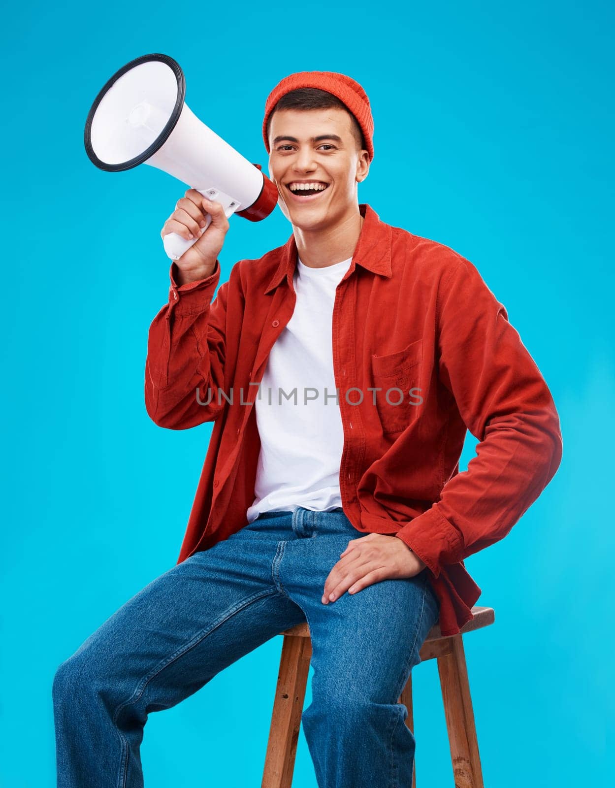 Portrait, megaphone and young man in a studio for an announcement or speech for a rally. Happy, smile and male activist on stool with bullhorn for loud communication isolated by blue background