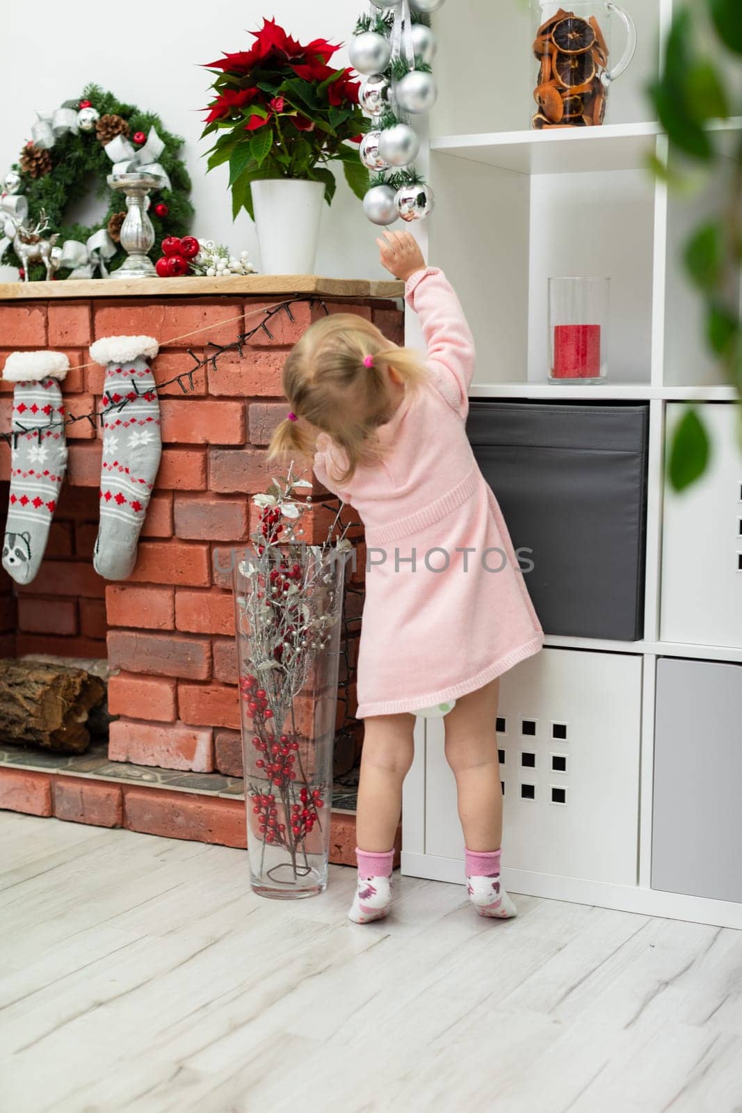 A little girl is standing by the fireplace extending her hand upward toward the silver baubles. The child is wearing a pink dress and standing with her back. The room is decorated with many Christmas decorations