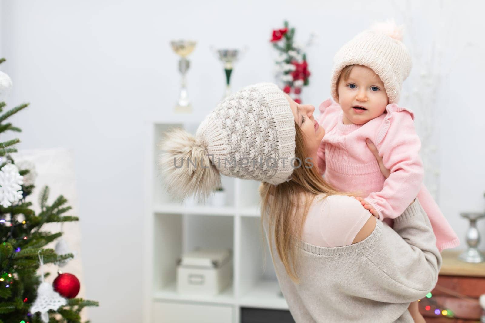 A mother carries her little daughter in her arms and smiles at her. The girl is wearing a pink dress and a cap. A fuzzy outline of a white bookcase can be seen in the background. A fragment of a Christmas tree can be seen next to the woman and her daughter.