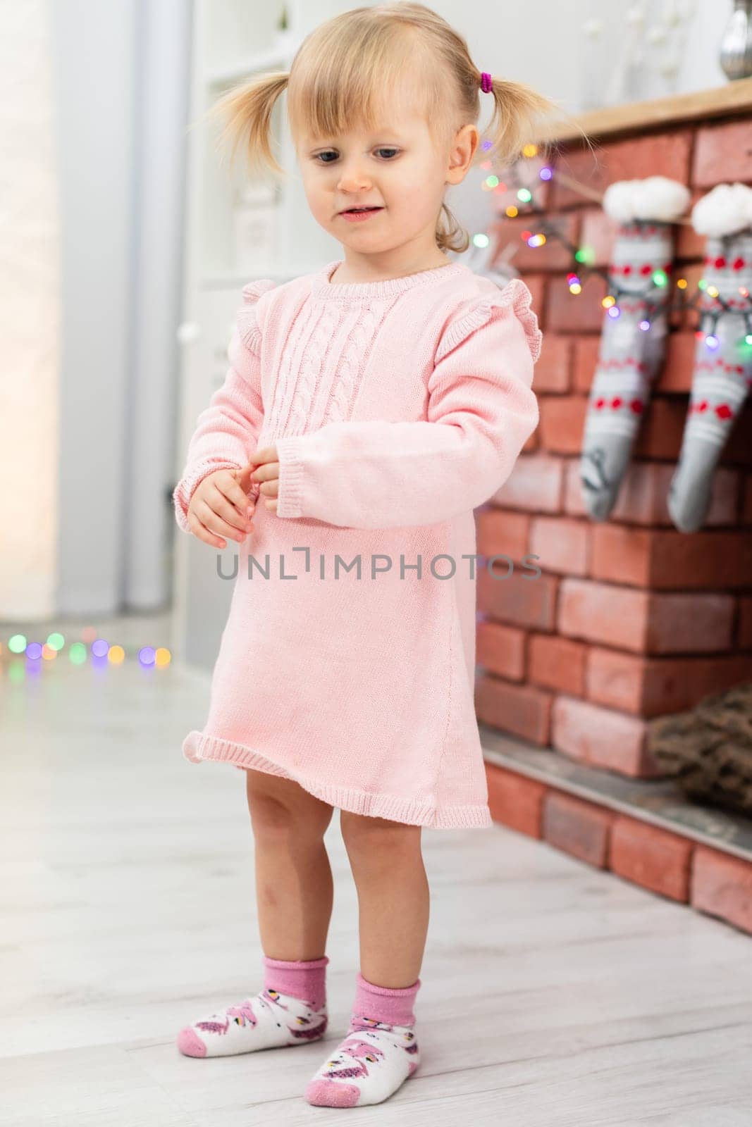 A tiny girl is standing in a room on a bright floor. The child is wearing a pink dress and colorful socks. The girl is standing by the fireplace, on which hang Christmas socks and colorful lights